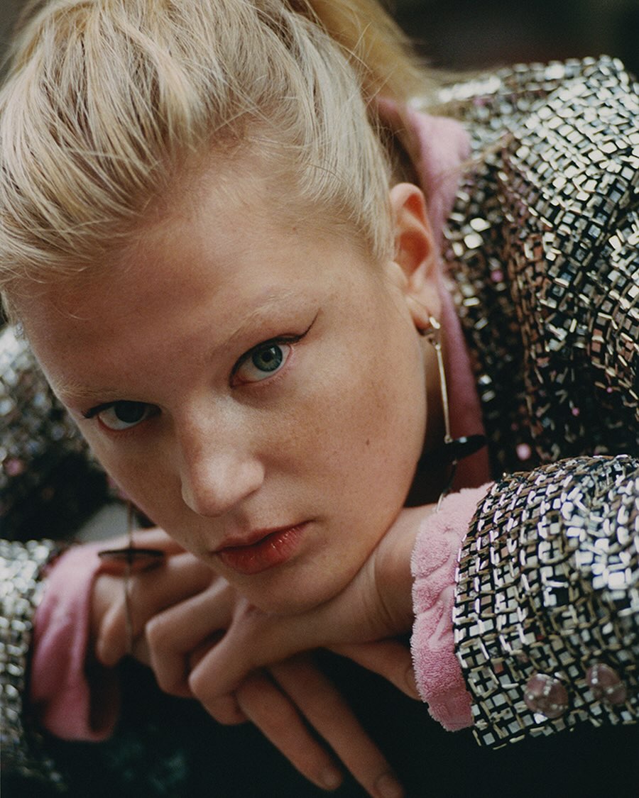Close-up portrait of a young blond woman with green eyes wearing a shimmering silver blazer, resting her face on her hands.