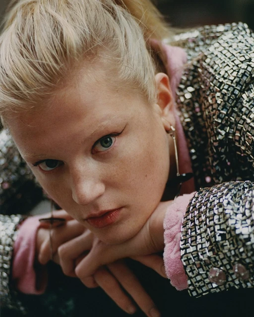 Close-up portrait of a young blond woman with green eyes wearing a shimmering silver blazer, resting her face on her hands.