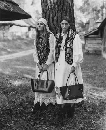 Two women in traditional clothing stand with leather tote bags in front of a tree. The black and white shot highlights historical fashion.