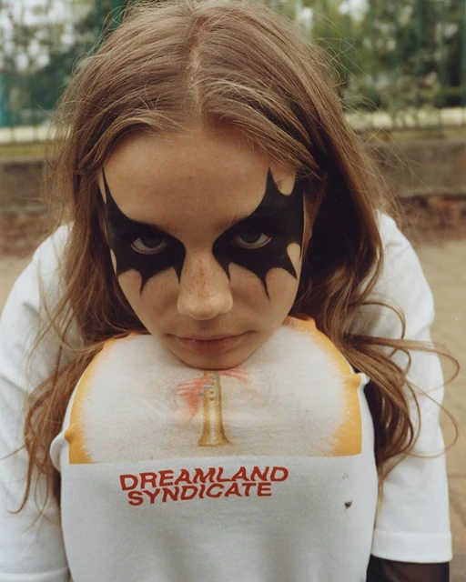 Young person with dramatic black eye makeup and a 'Dreamland Syndicate' t-shirt poses outdoors against a blurred greenery backdrop.