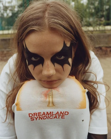 Young person with dramatic black eye makeup and a 'Dreamland Syndicate' t-shirt poses outdoors against a blurred greenery backdrop.