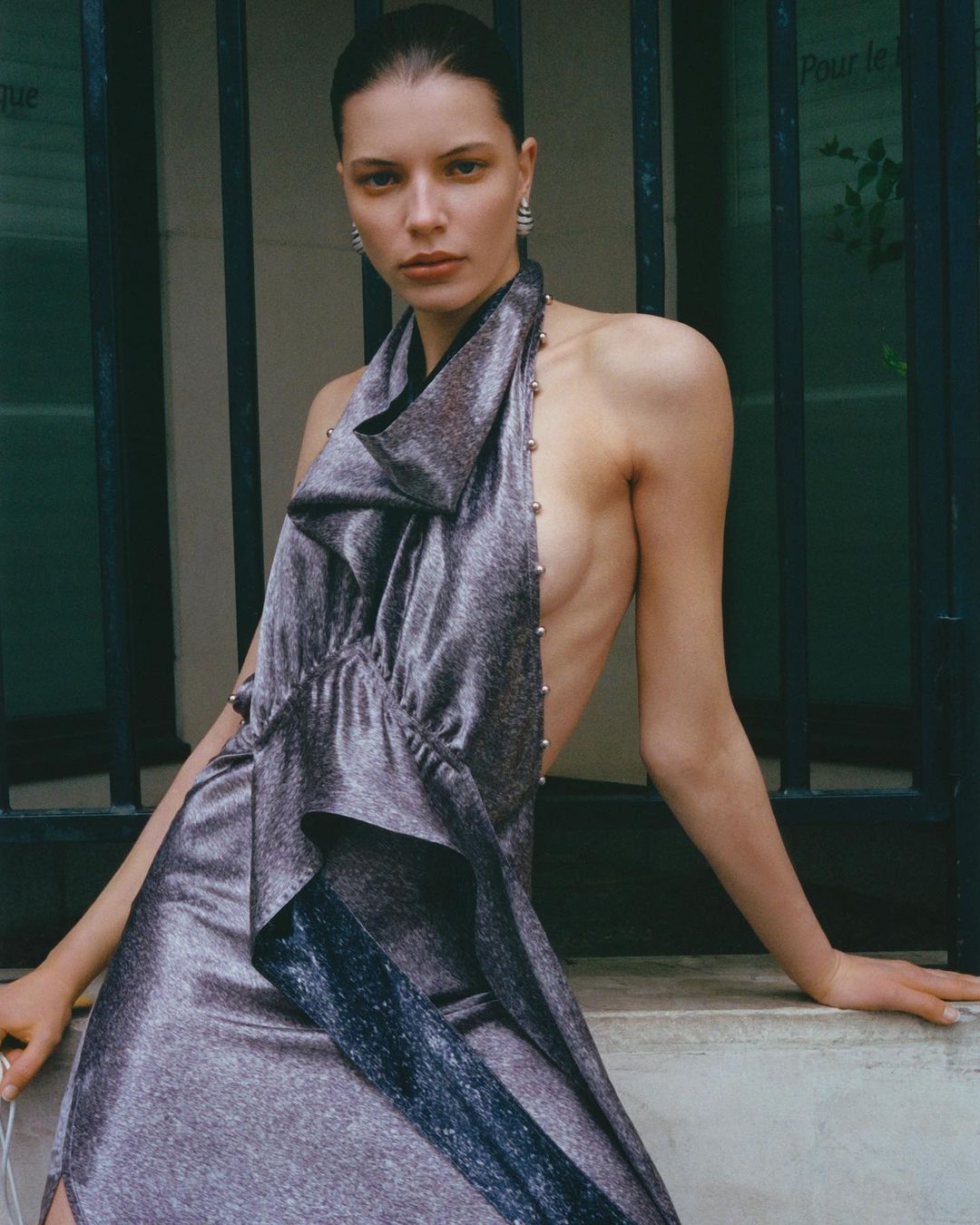 Adult woman poses in silver dress, against an outdoor stone window ledge. Fashion editorial shot with natural light.
