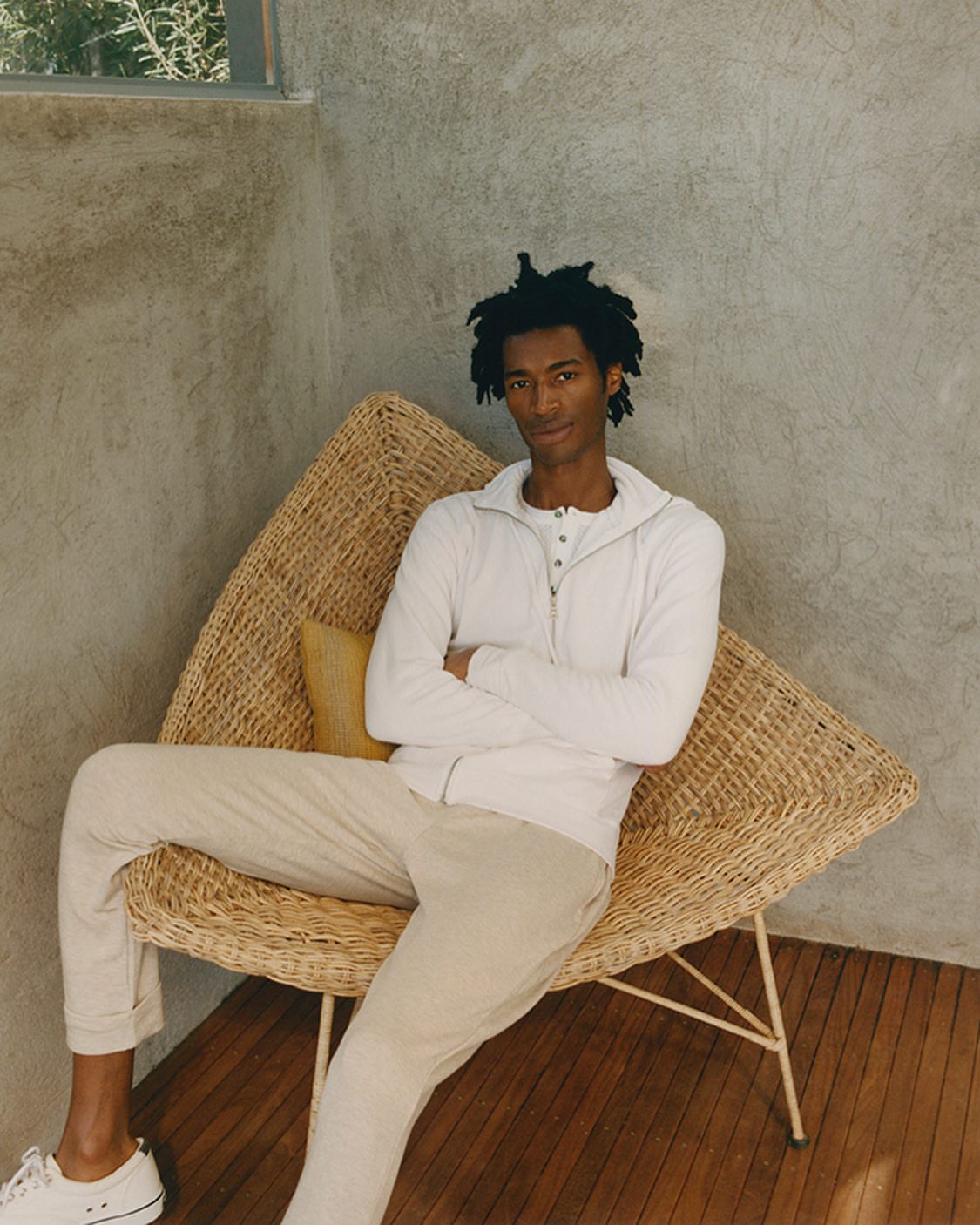 An adult male sits cross-legged in a wicker chair on a wood floor in front of a neutral wall, wearing a white jacket and light-colored pants.