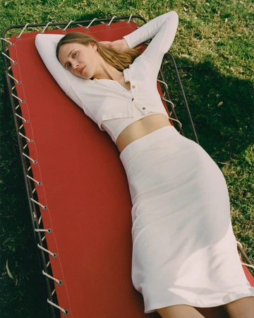 Woman in white crop top and skirt lounging on a red cot on green grass, angled eye-level shot, soft lighting, neutral color grading.