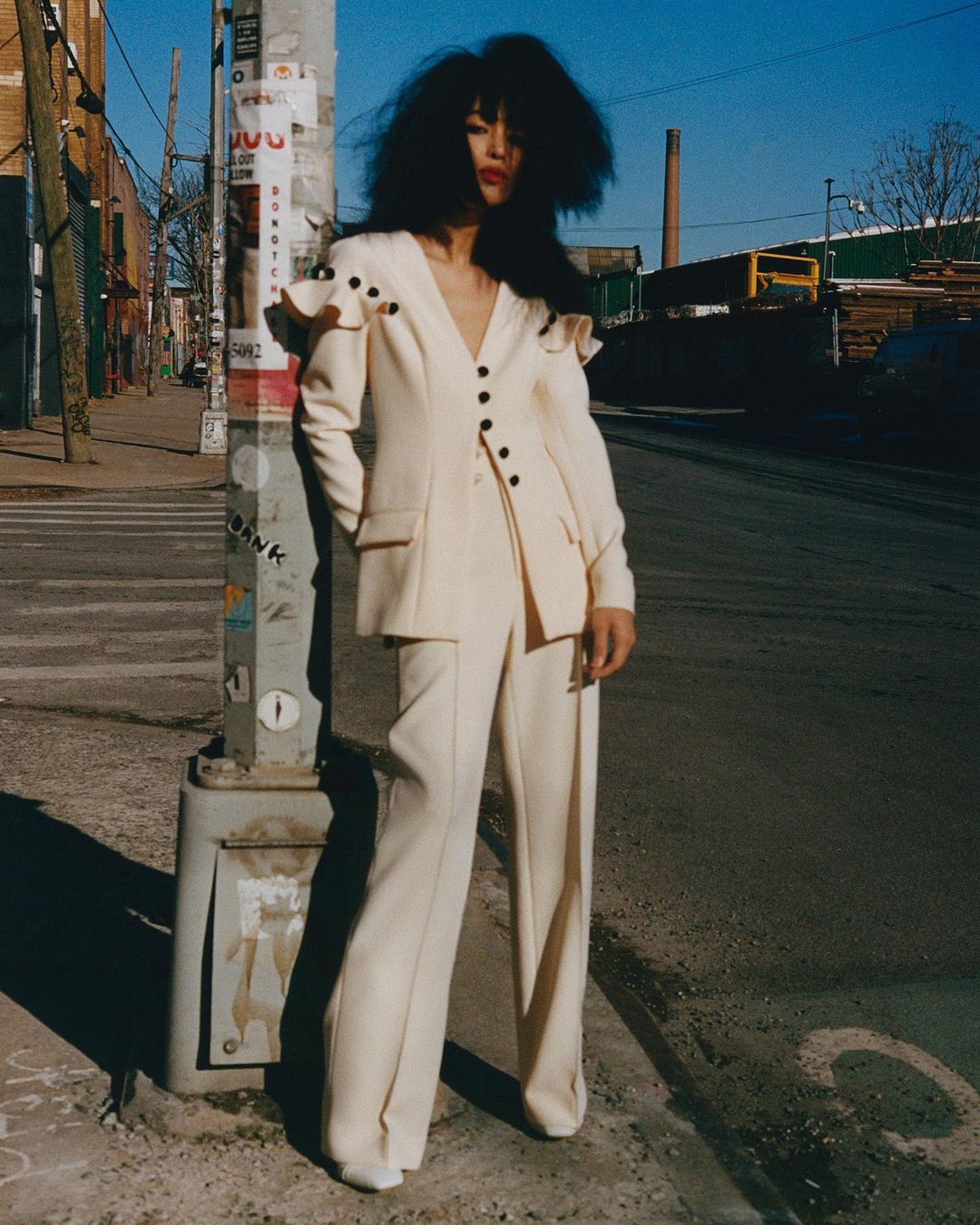 A woman in a cream suit stands on a city street next to a utility pole on a sunny day, showcasing high-fashion in an urban setting.