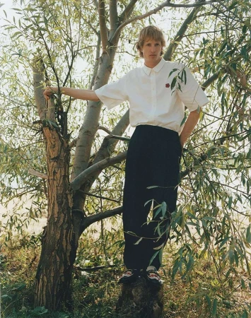 A blond-haired model wearing a white short-sleeved shirt and black pants stands atop a tree stump in a lush, sunlit forest setting.