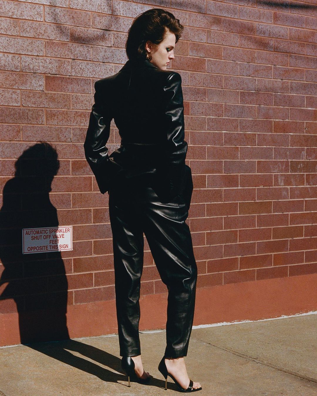 Woman in a black leather suit posing against a brick wall. She's wearing high heels and her gaze is directed slightly downward.