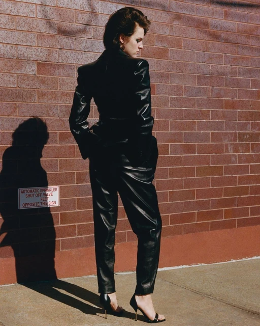 Woman in a black leather suit posing against a brick wall. She's wearing high heels and her gaze is directed slightly downward.