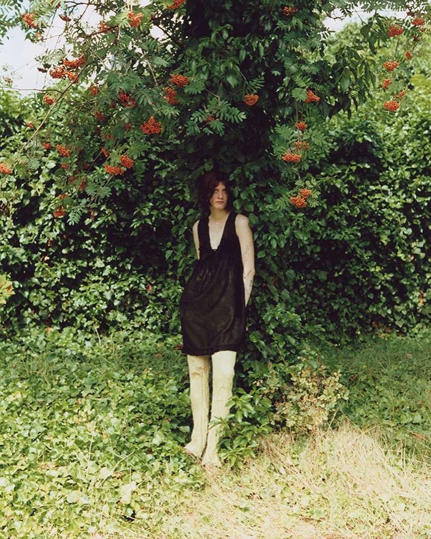 A woman in a black dress stands beneath a tree with red berries in a lush, verdant outdoor setting, captured in a medium shot.