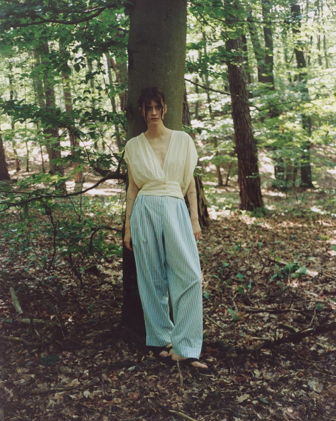 A woman in a light-yellow top and striped pants stands near a tree in a sunlit forest. Fashion shot with dappled light.