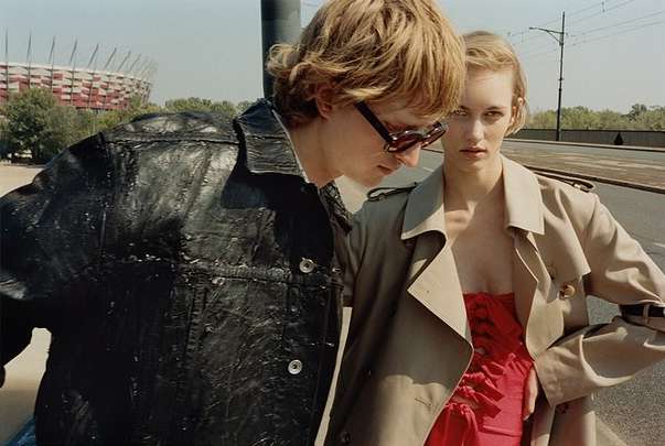 Two fashion models pose outdoors on a sunny day; one in a trench coat and the other in a black jacket near an urban landscape and stadium.