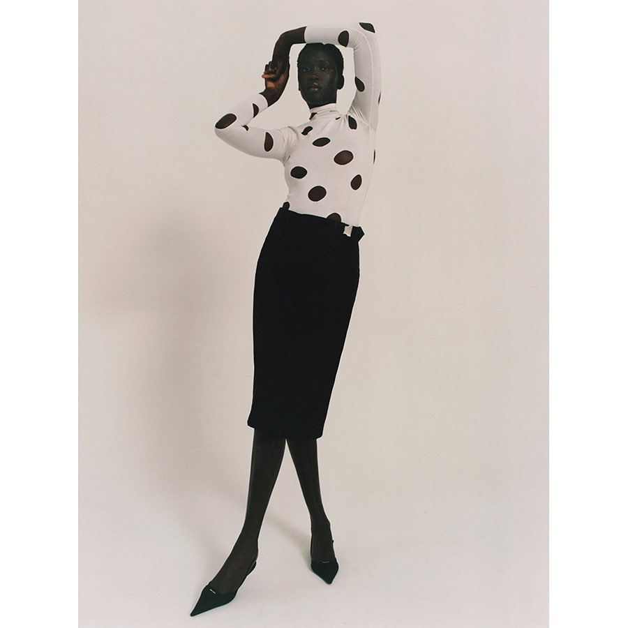 Fashion shot of a Black female model wearing a white turtleneck with black polka dots, a black pencil skirt, and black heels in studio.