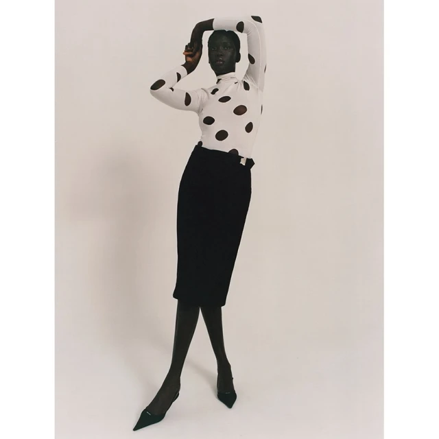 Fashion shot of a Black female model wearing a white turtleneck with black polka dots, a black pencil skirt, and black heels in studio.