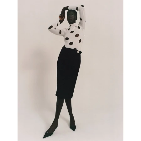 Fashion shot of a Black female model wearing a white turtleneck with black polka dots, a black pencil skirt, and black heels in studio.