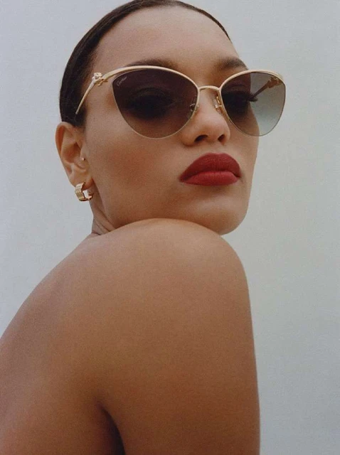 Adult female model poses in a close-up shot wearing sunglasses and gold hoop earrings against a plain light background in a beauty-style image.