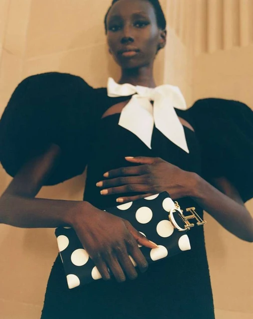 Elegant woman in black dress with white bow poses with a black and white polka dot clutch indoors against a neutral background.