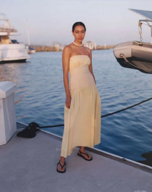 Elegant adult female in a long, strapless dress standing on a dock near boats, under soft natural light, and wearing flat sandals.
