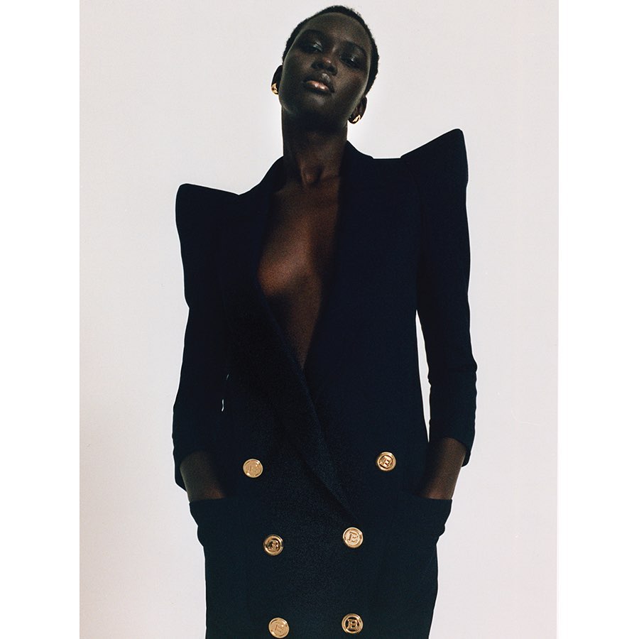 Portrait of a Black female model in a black dress with gold buttons against a white backdrop in a fashion editorial photograph.