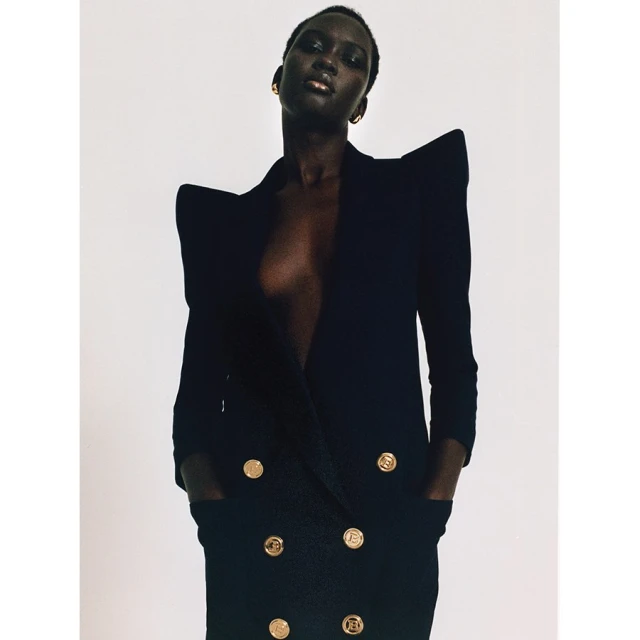 Portrait of a Black female model in a black dress with gold buttons against a white backdrop in a fashion editorial photograph.