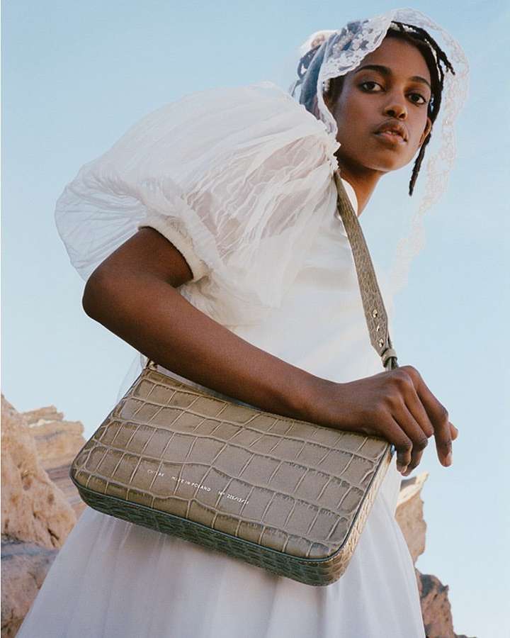 Woman in white dress poses with a textured handbag outdoors against a blue sky. Fashion portrait showcasing accessories.
