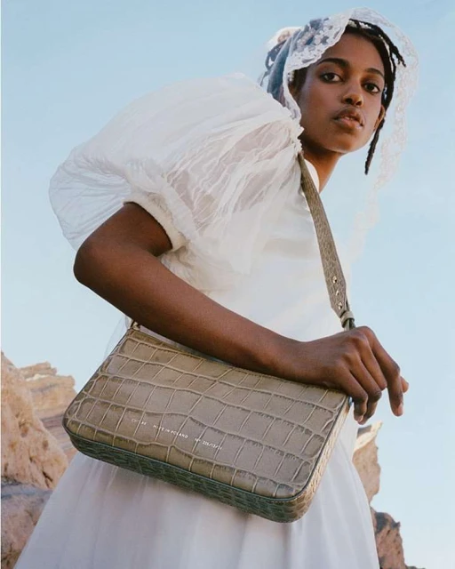 Woman in white dress poses with a textured handbag outdoors against a blue sky. Fashion portrait showcasing accessories.