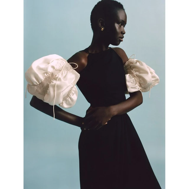 A sophisticated fashion portrait of a black female model in a black dress with voluminous white statement sleeves against a light blue backdrop.