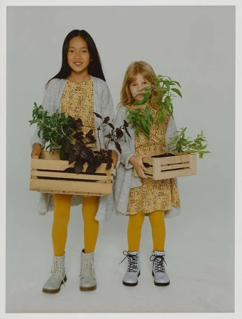 Two young girls in matching dresses and yellow tights holding wooden crates with plants in a photo studio setting with a soft light.