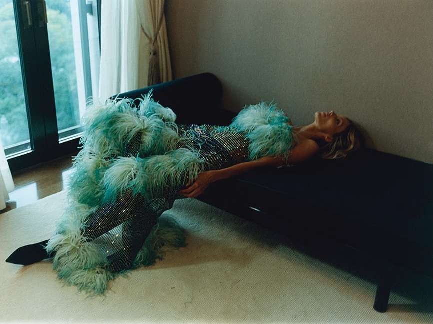 Woman in sparkling dress and feathered coat lies on a black chaise lounge, creating a luxurious and fashionable editorial image indoors.