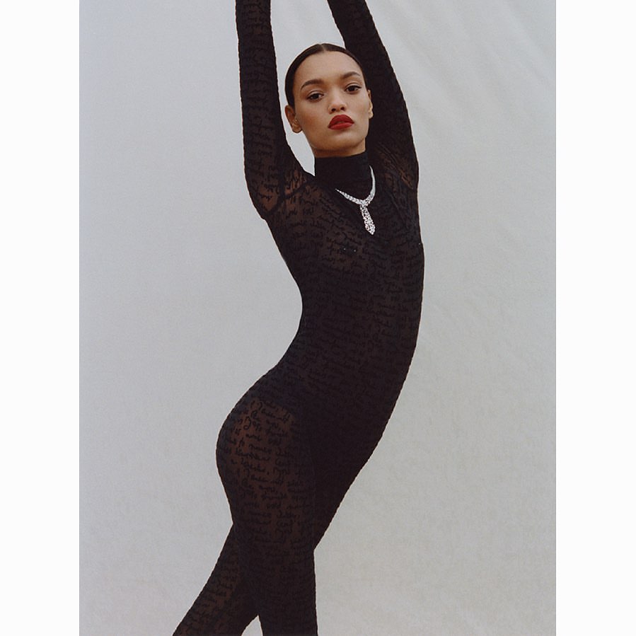 Woman in long-sleeved black patterned bodysuit poses with arms raised, wearing a diamond necklace, against a light grey backdrop.