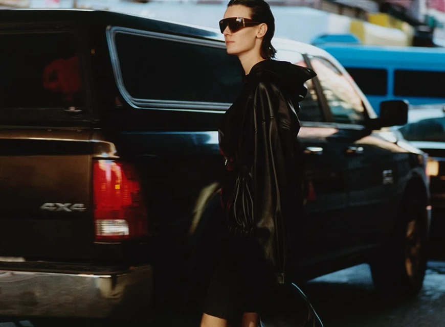 A woman in sunglasses and a leather jacket walks past a black pickup truck in an urban setting with neutral, muted color tones.