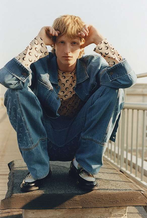 Man with blond hair squats on rooftop, wearing denim jacket and jeans with crescent moon print top and black loafers in daylight.