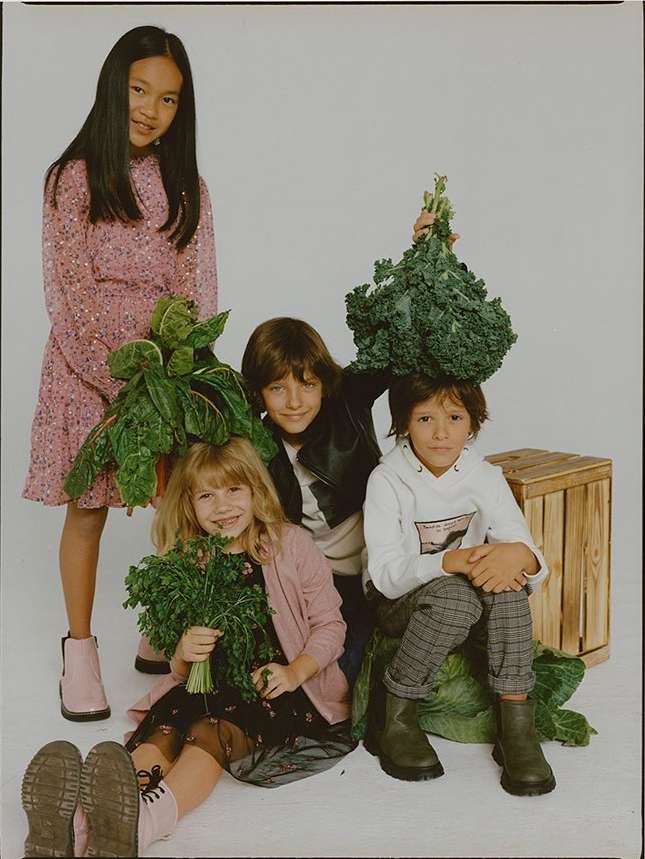 Four children pose with leafy greens against a white backdrop, one child standing and three seated near a crate and dark footwear.