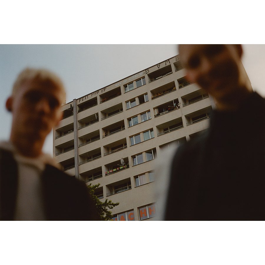 Two young men stand in front of a tall apartment building, the architecture dominates the frame with its many windows and orange signage.