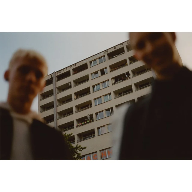 Two young men stand in front of a tall apartment building, the architecture dominates the frame with its many windows and orange signage.