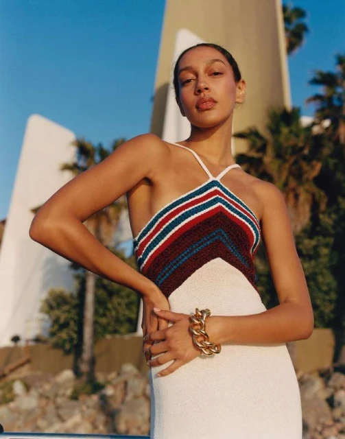 Brunette woman in halter dress poses outdoors with a building and palm trees. Gold chain bracelet adds a striking touch.