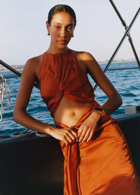 A woman poses in a burnt-orange skirt and halter top on a boat, leaning on the railing with a sunny sea and horizon in the background.