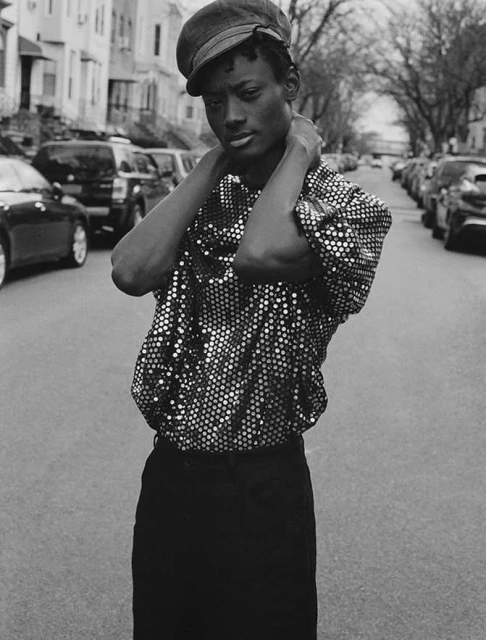 A black and white portrait of a male model wearing a sequined shirt and hat standing on a city street with cars and buildings.
