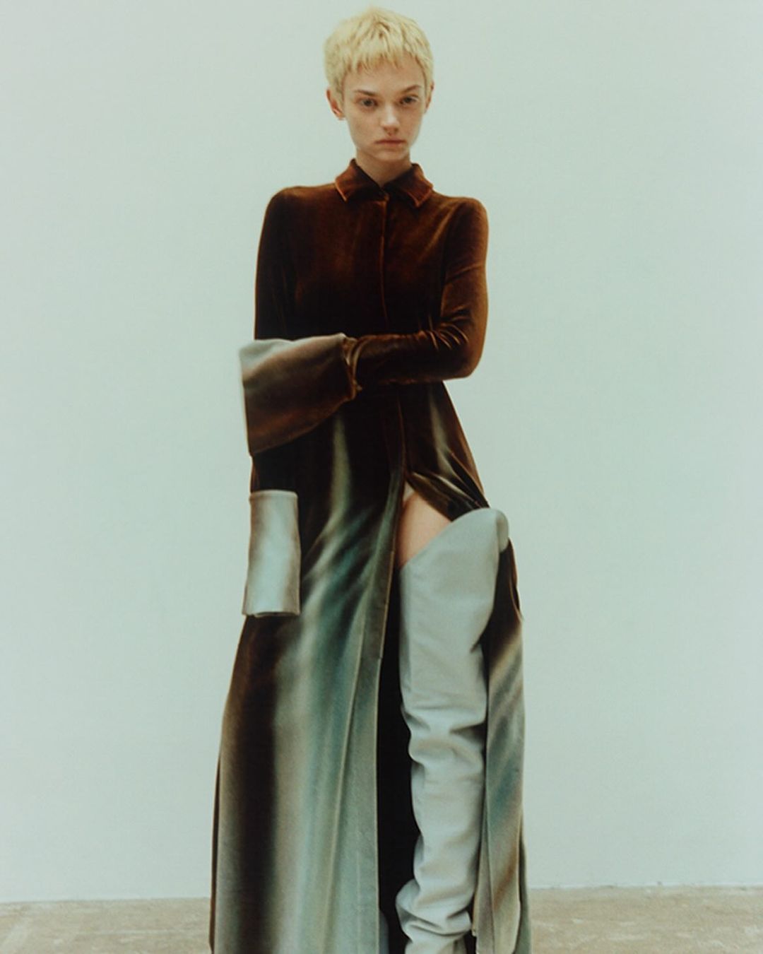 A woman poses in a long brown velvet dress with a thigh-high slit and high gray boots against a neutral background in a fashion shot.