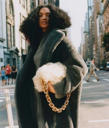 Model with voluminous dark hair wearing a charcoal grey oversized teddy coat carrying a fluffy white handbag with a chunky gold chain