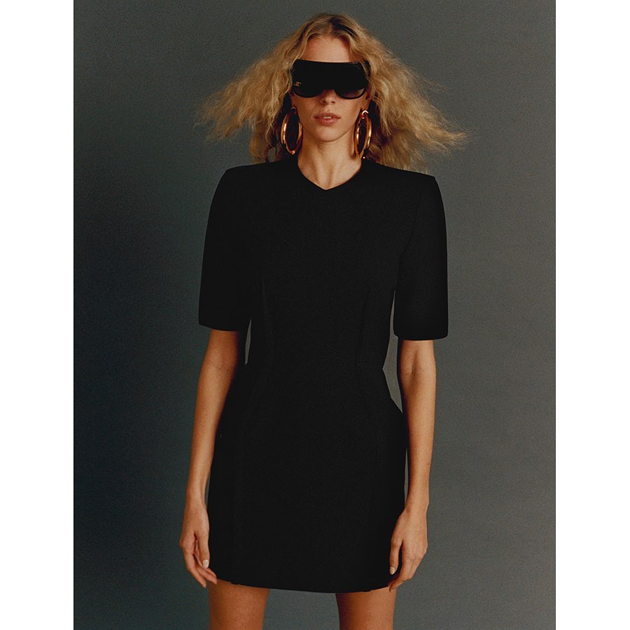 A woman in a black dress stands against a gray background, wearing futuristic sunglasses and hoop earrings for a fashion-forward image.