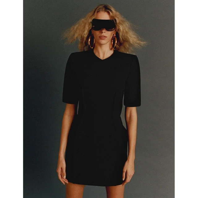 A woman in a black dress stands against a gray background, wearing futuristic sunglasses and hoop earrings for a fashion-forward image.