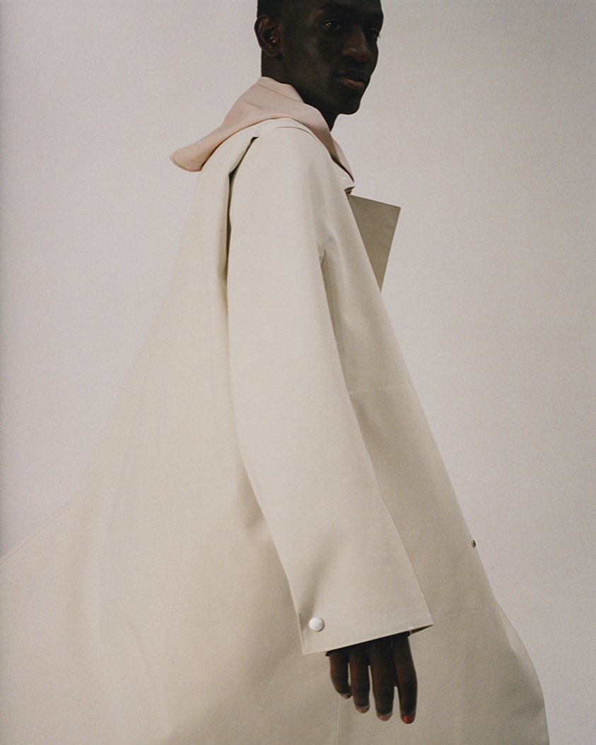 Studio shot of a black male model wearing a beige overcoat and light pink hoodie, gazing off camera with a neutral expression.