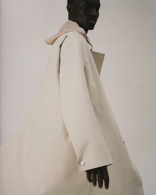 Studio shot of a black male model wearing a beige overcoat and light pink hoodie, gazing off camera with a neutral expression.