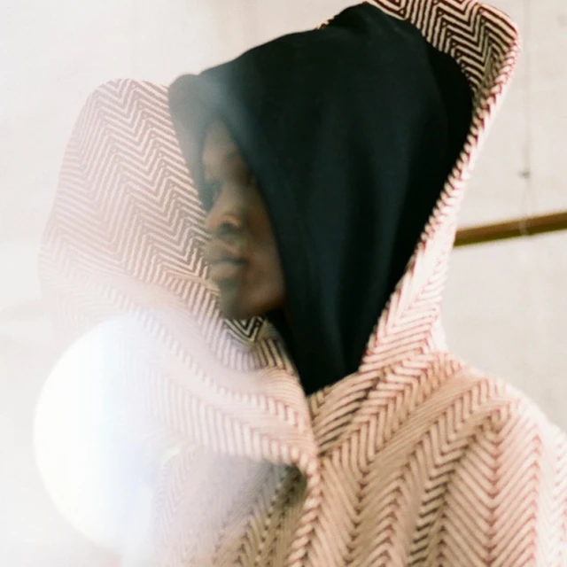 Fashion shot of a person with dark skin in a hooded jacket with a geometric pattern, side profile with soft lighting and a muted color palette.
