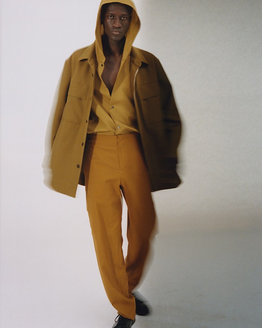 Fashionable adult male in an unbuttoned yellow shirt and mustard pants stands in a photo studio with his hands at his sides.