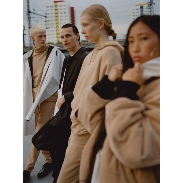 Four young adults in stylish beige and black streetwear pose outdoors against an urban backdrop of buildings and cloudy sky.