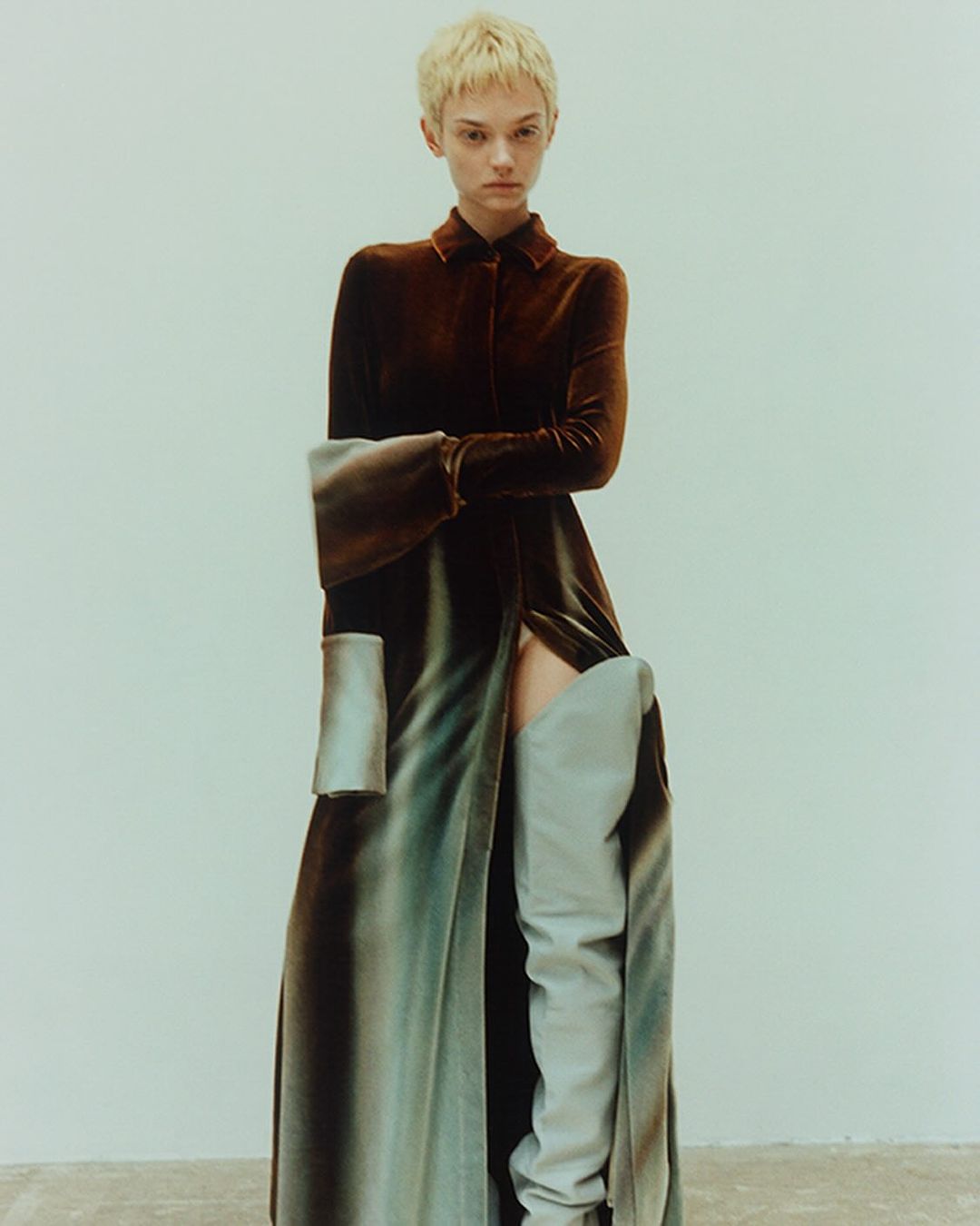 A woman with short blonde hair poses in a long, dark brown dress with long sleeves and a slit, paired with light blue thigh-high boots.
