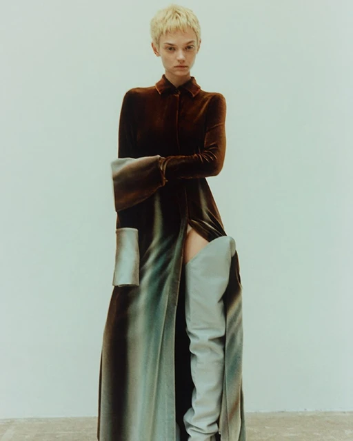 A woman with short blonde hair poses in a long, dark brown dress with long sleeves and a slit, paired with light blue thigh-high boots.