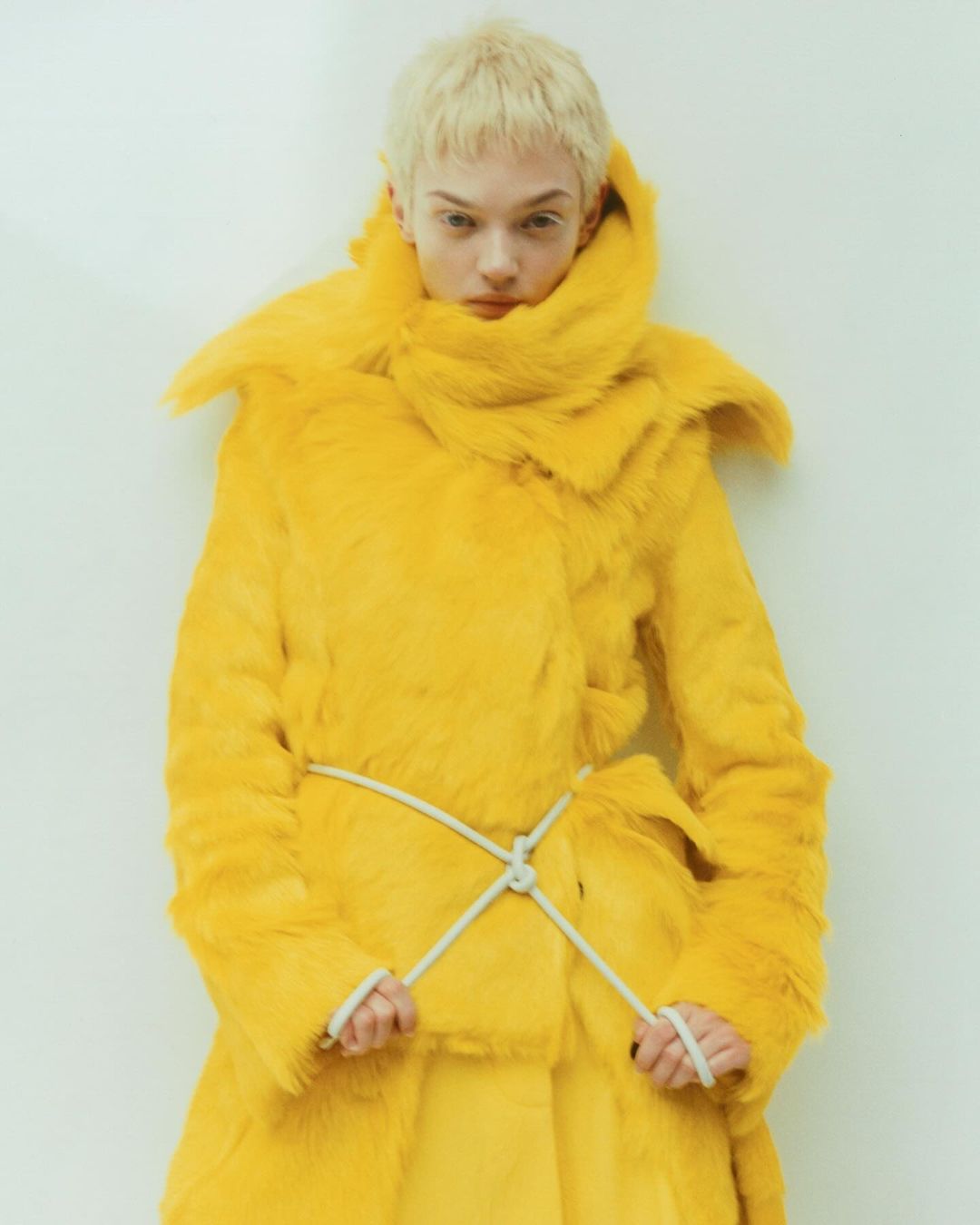 Fashionable young woman with short blonde hair in a bright yellow faux fur coat with a rope belt posing against a white wall.