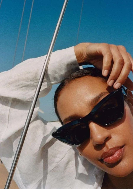 Woman in a white linen shirt and black sunglasses poses on a yacht, against a clear blue sky on a sunny day.